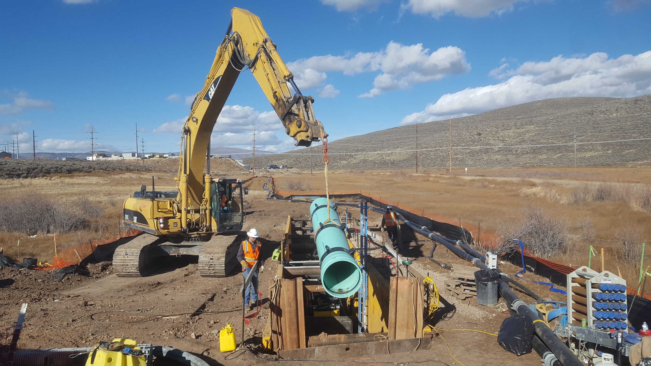 Excavator lowering large blue pipe on site of Silver Creek Trunkline Sewer Rehabilitation project as a construction worker watches.