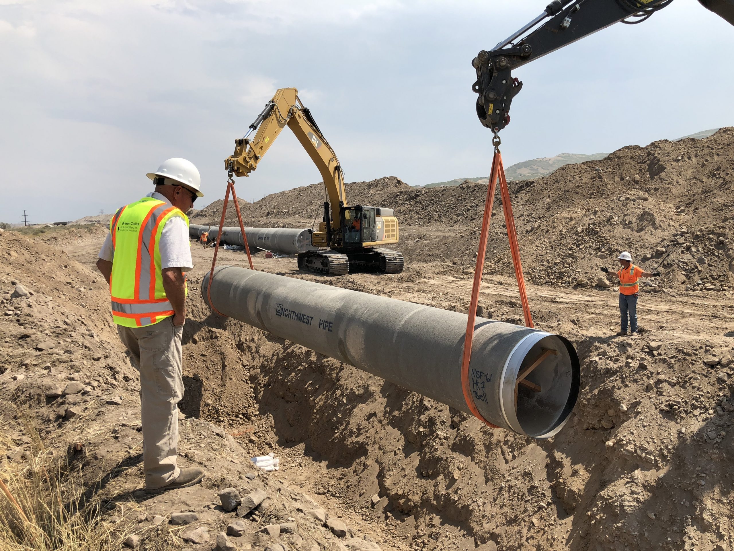 Construction worker observing large metal pipe being lowered into the ground by cranes.