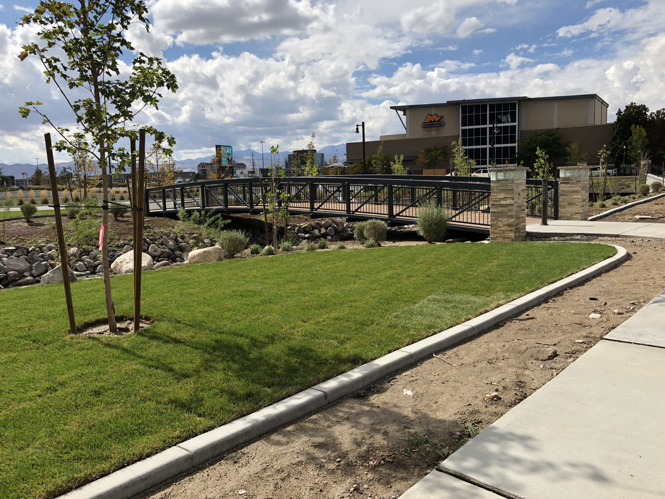 Pedestrian bridge over Dry Creek channel with surrounding grass, buildings, and sidewalks.