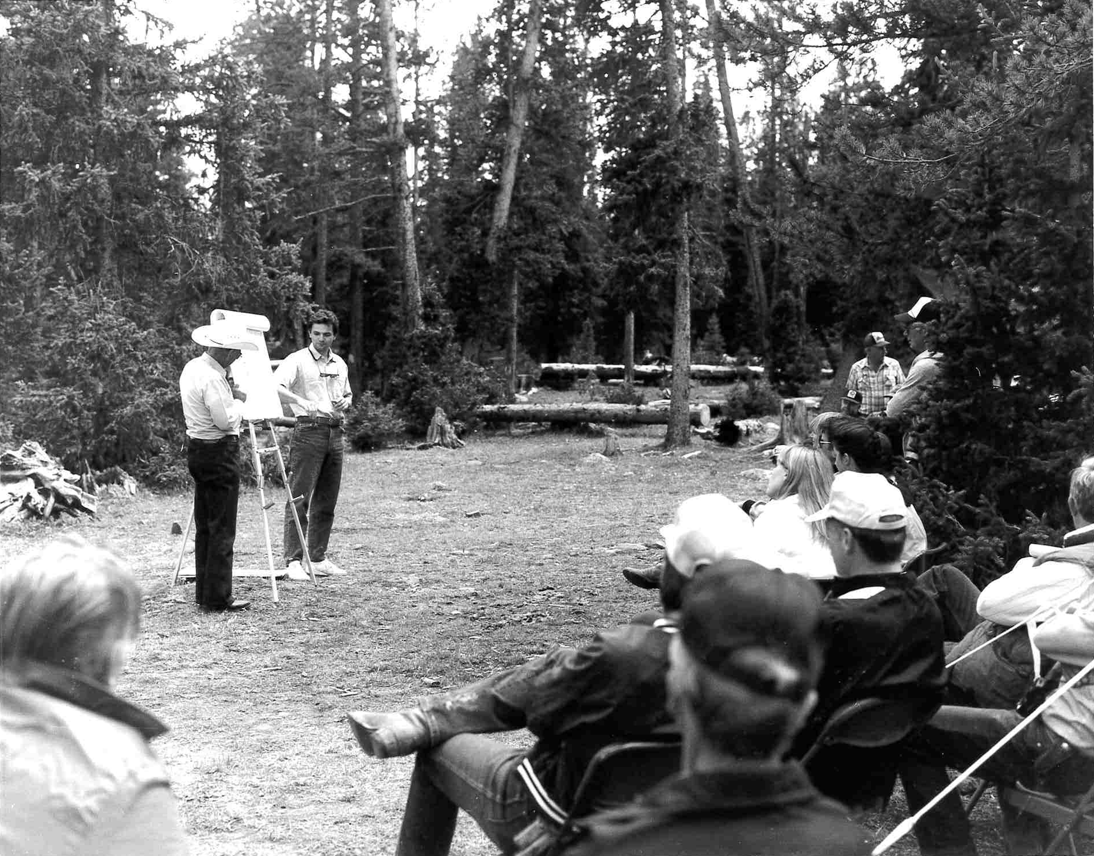 People gathered in forest discussing materials displayed on a stand